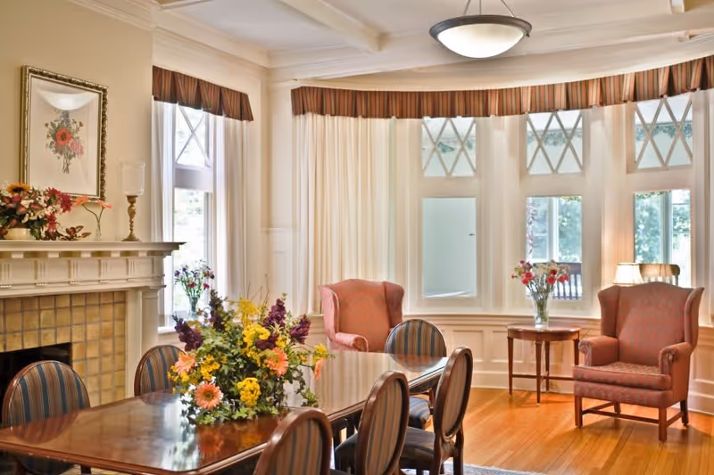 A bright and elegant living room with a wooden dining table surrounded by six striped chairs. Two upholstered armchairs are positioned near large windows with white curtains and valances. A fireplace with a floral painting above it and a vase of flowers on the mantel is visible on the left side. The room has hardwood floors and a ceiling light fixture.
