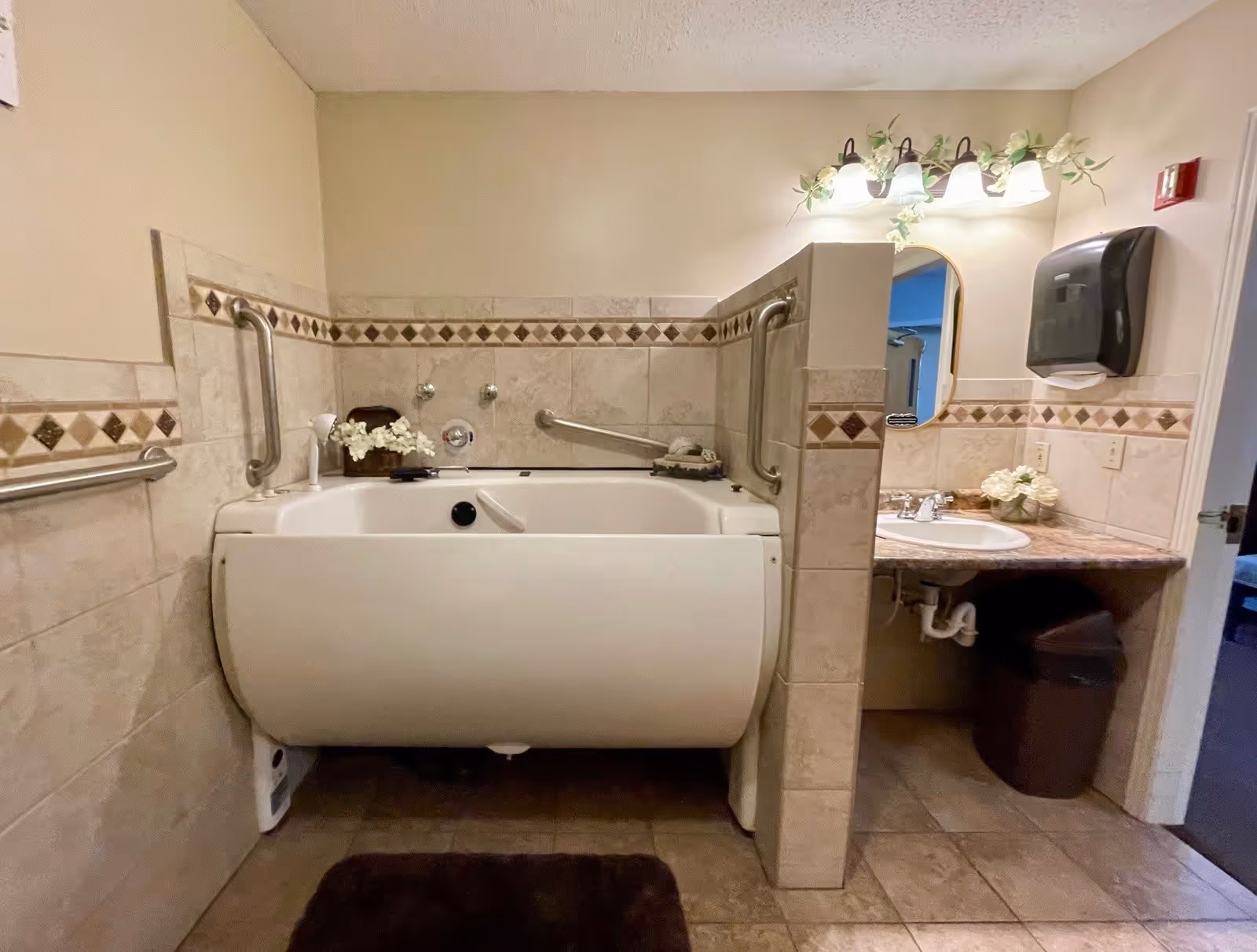 A bathroom with a walk-in bathtub featuring grab bars on the walls for safety. The walls are tiled with a decorative border. To the right, there is a sink with a marble countertop, a mirror, a light fixture with four bulbs, a paper towel dispenser, and a trash bin underneath the sink. The floor is tiled and there is a dark brown bath mat in front of the tub.