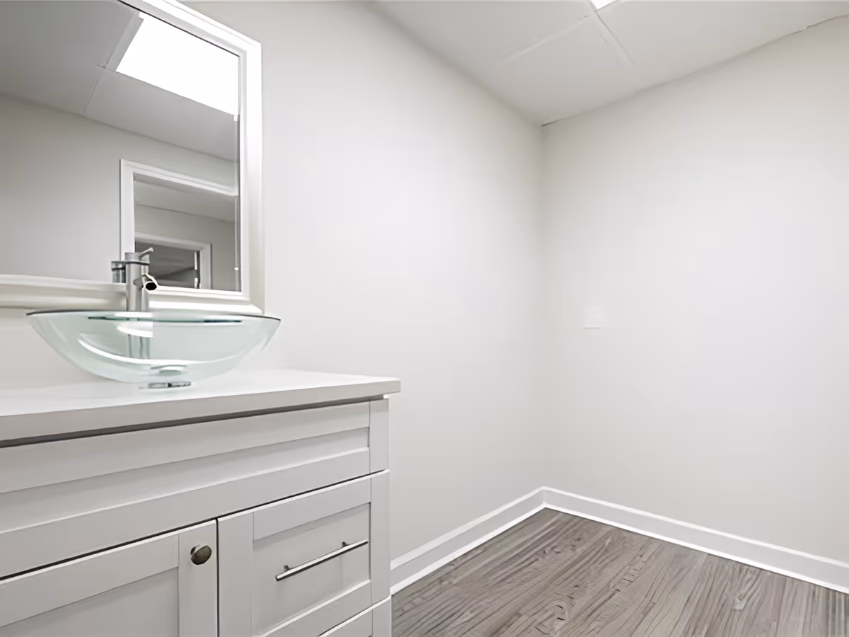 A modern bathroom corner featuring a white vanity with a glass vessel sink and a chrome faucet. Above the vanity is a rectangular mirror with a white frame. The walls are painted white, and the floor has a wood-like finish.