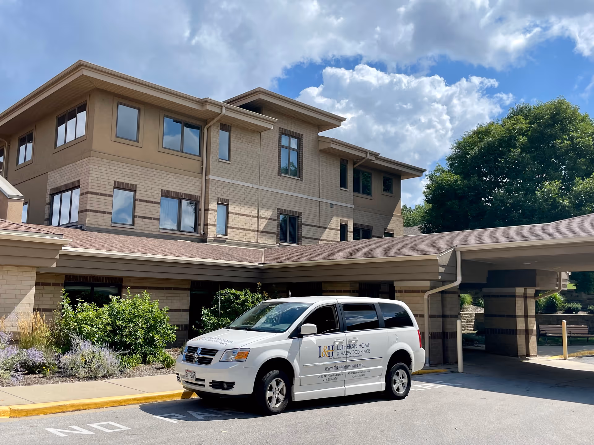 Exterior view of a multi-story senior living facility building with beige brick and tan siding. A white van with the logo and text 'Lutheran Home & Harwood Place' is parked in front near a covered entrance. There are green bushes and trees around the building under a partly cloudy blue sky.