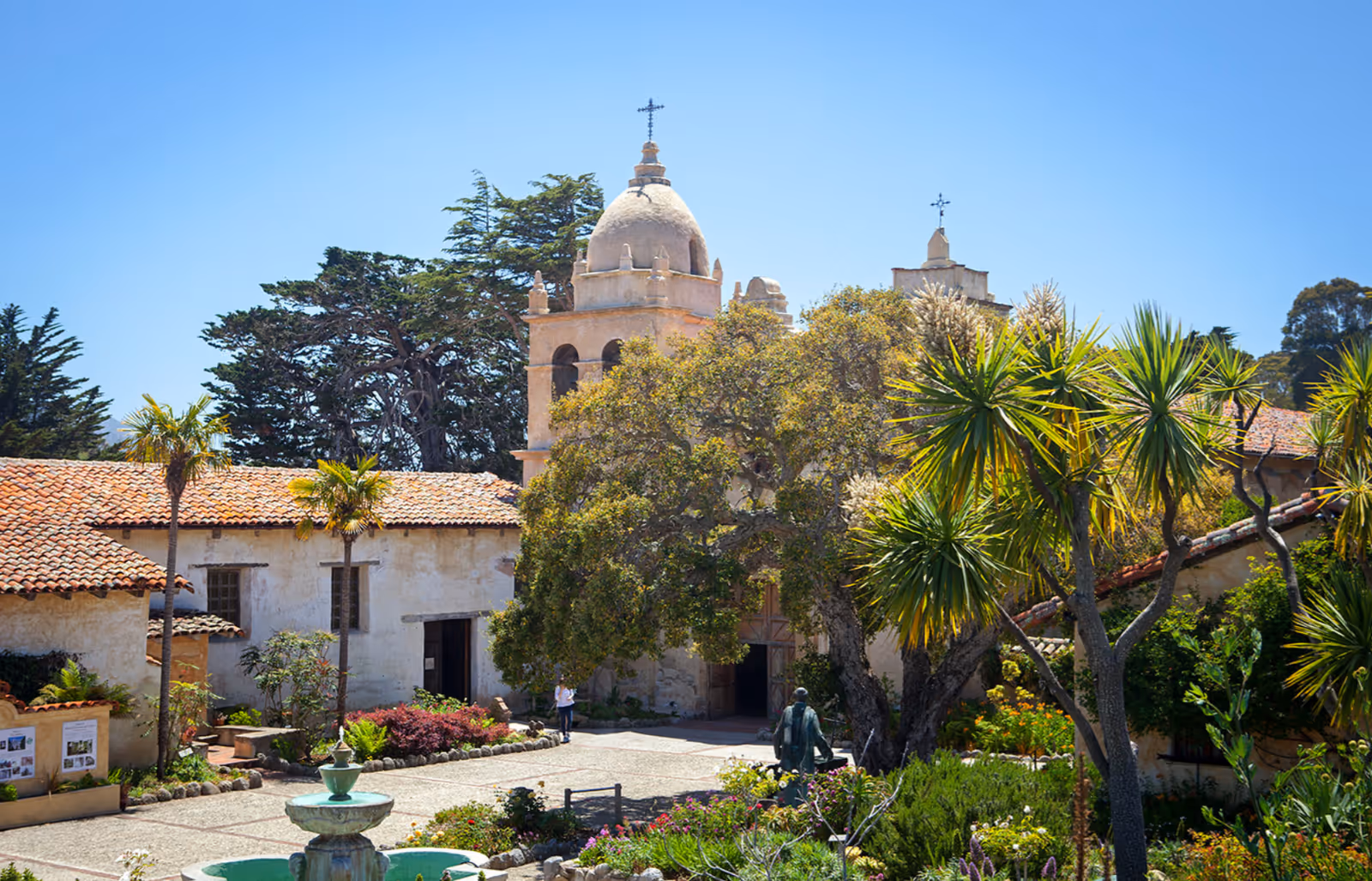 A sunny courtyard with a fountain in the center, surrounded by lush greenery and palm trees. In the background, there is a historic building with a bell tower topped with a cross, featuring a tiled roof and arched windows.