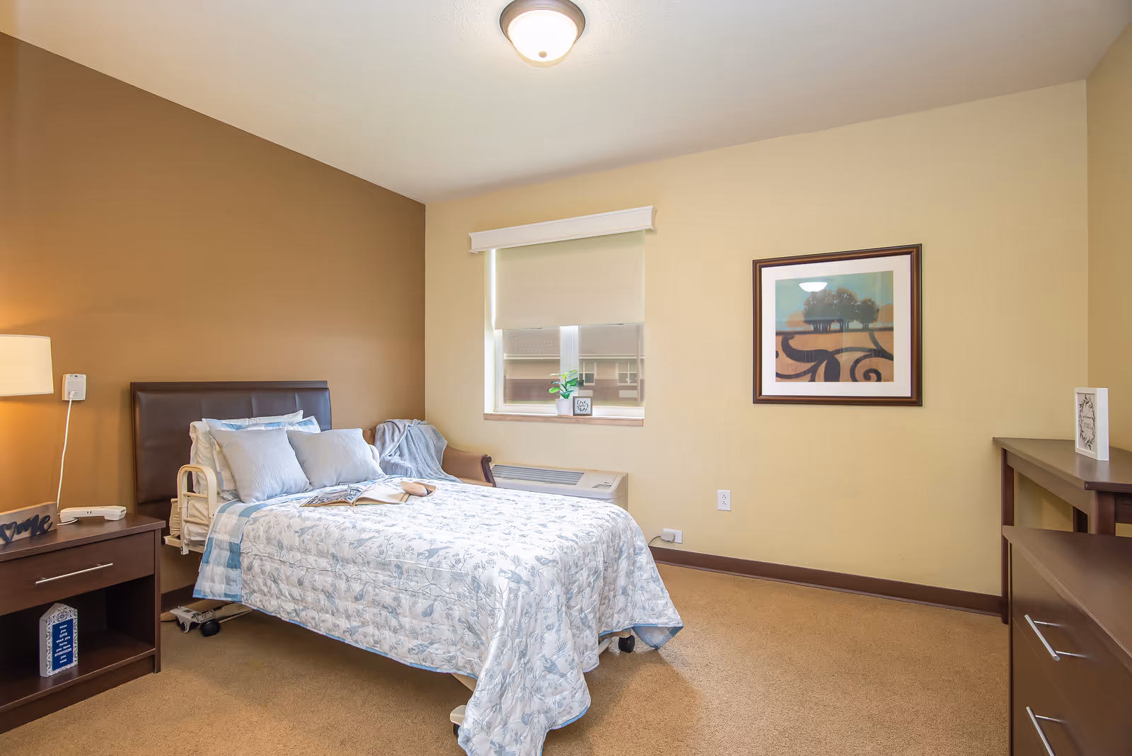 A tidy single bedroom with a bed, nightstand and lamp, a window, framed wall art, and simple furnishings.