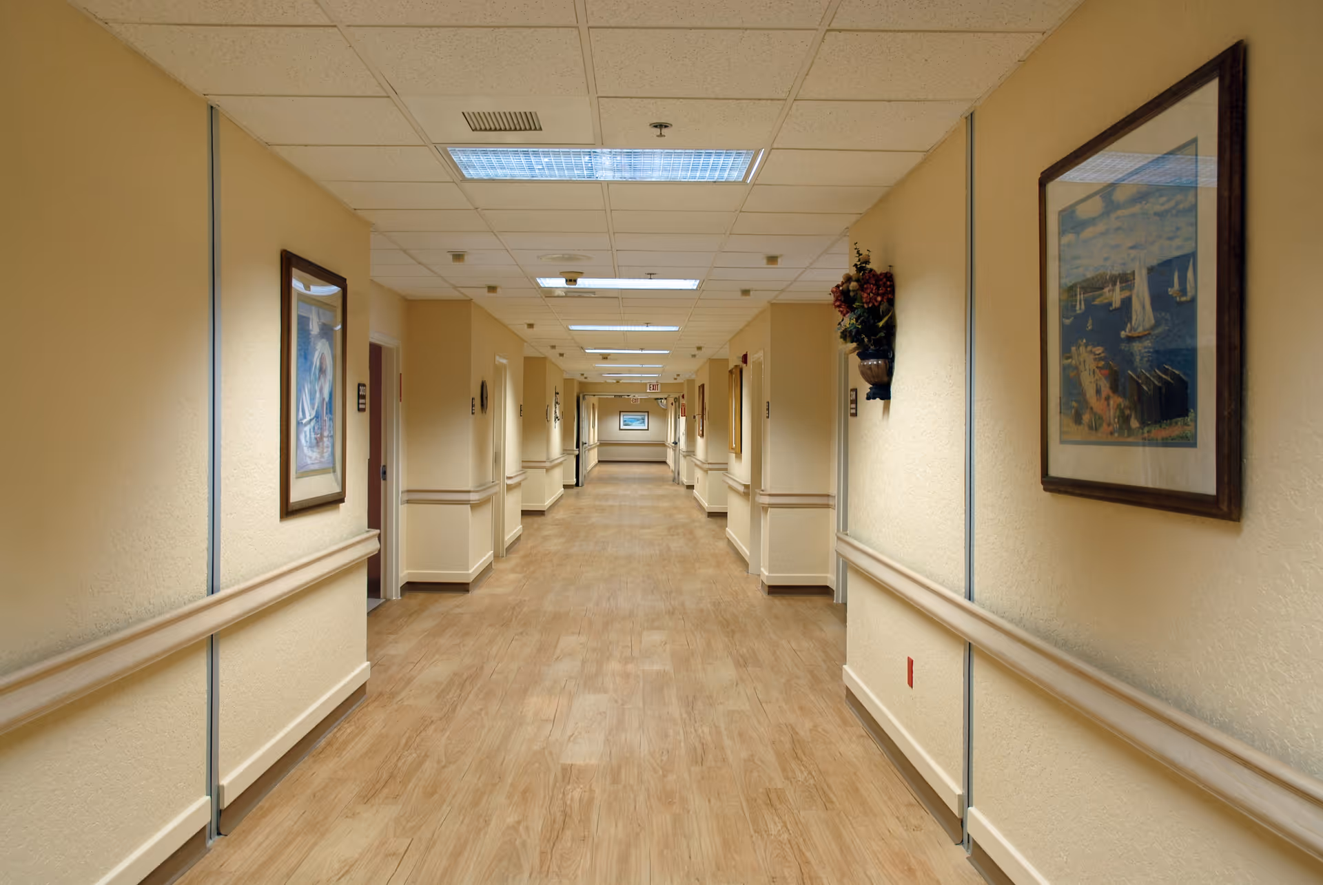 A long, well-lit hallway in a senior living facility with light beige walls, wooden handrails on both sides, framed paintings hanging on the walls, and a wood-patterned floor. The ceiling has recessed fluorescent lighting panels.