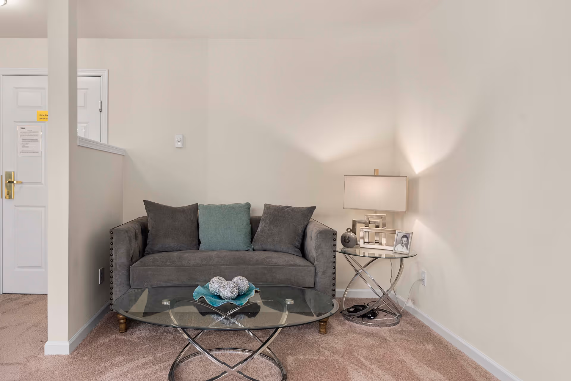 Small living area with a gray loveseat, glass coffee table, and a side table with a lamp and framed photo against neutral walls.