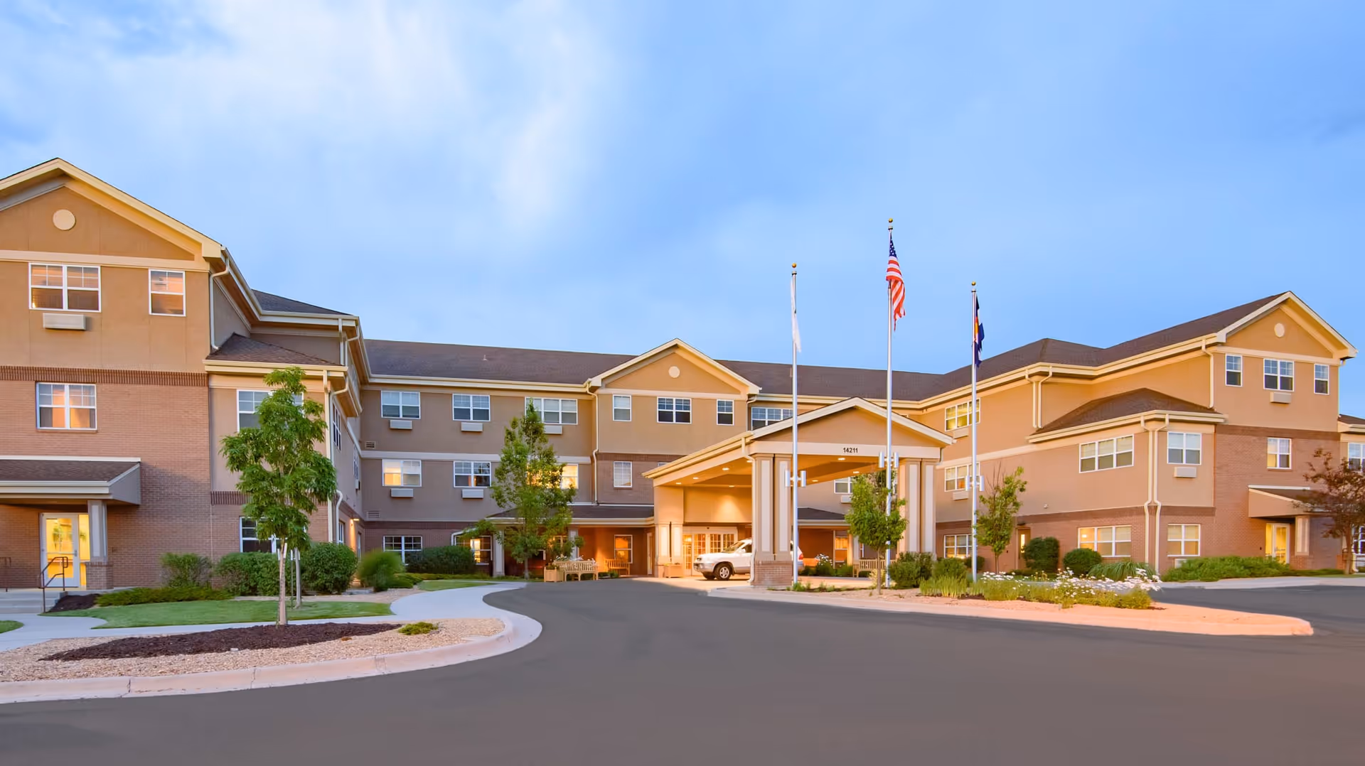 Exterior view of a large senior living facility building with three stories, beige and brown walls, multiple windows, and a covered entrance with three flagpoles displaying flags. The driveway and landscaped areas with trees and shrubs are visible in front of the building under a clear blue sky.