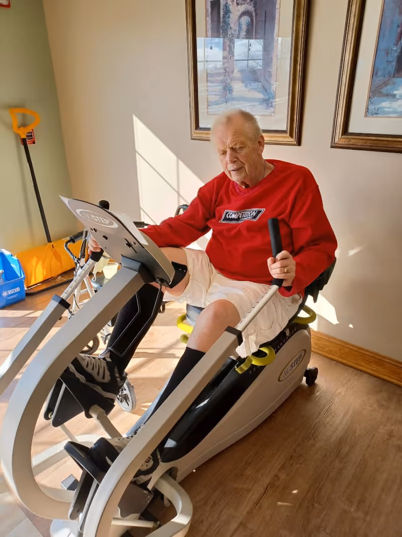 An elderly man wearing a red sweatshirt and white shorts is exercising on a NuStep recumbent cross trainer machine in a sunlit room with wooden floors and framed artwork on the wall behind him.