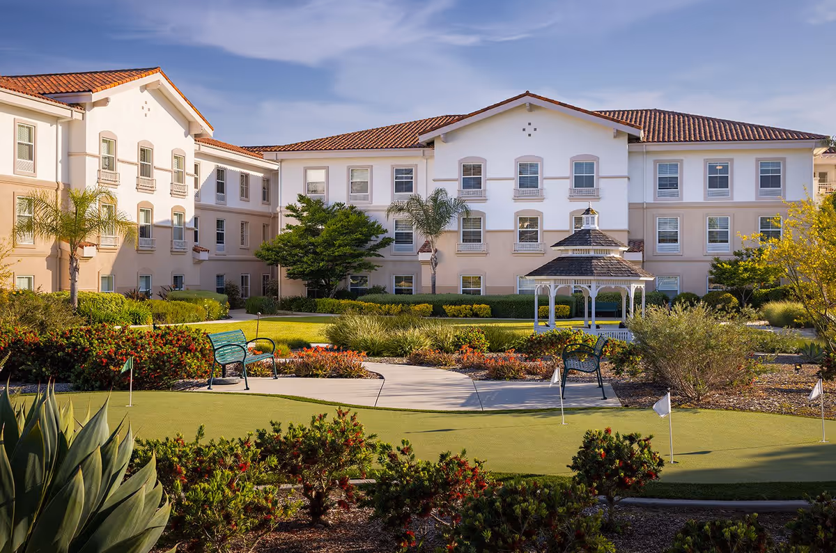 Exterior view of Belmont Village Senior Living Sabre Springs showing a three-story building with a red-tiled roof, surrounded by landscaped gardens, palm trees, and a putting green with benches and a white gazebo in the courtyard.