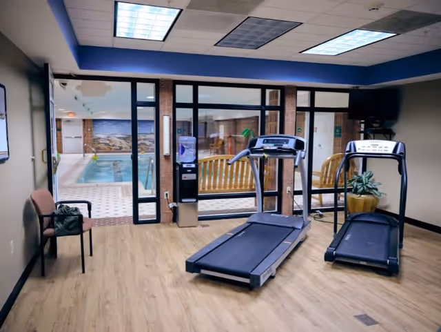 Indoor fitness room with two treadmills on a wooden floor, a chair with a bag on it, a water dispenser, and a large window looking into an indoor swimming pool area with a bench and tiled floor.