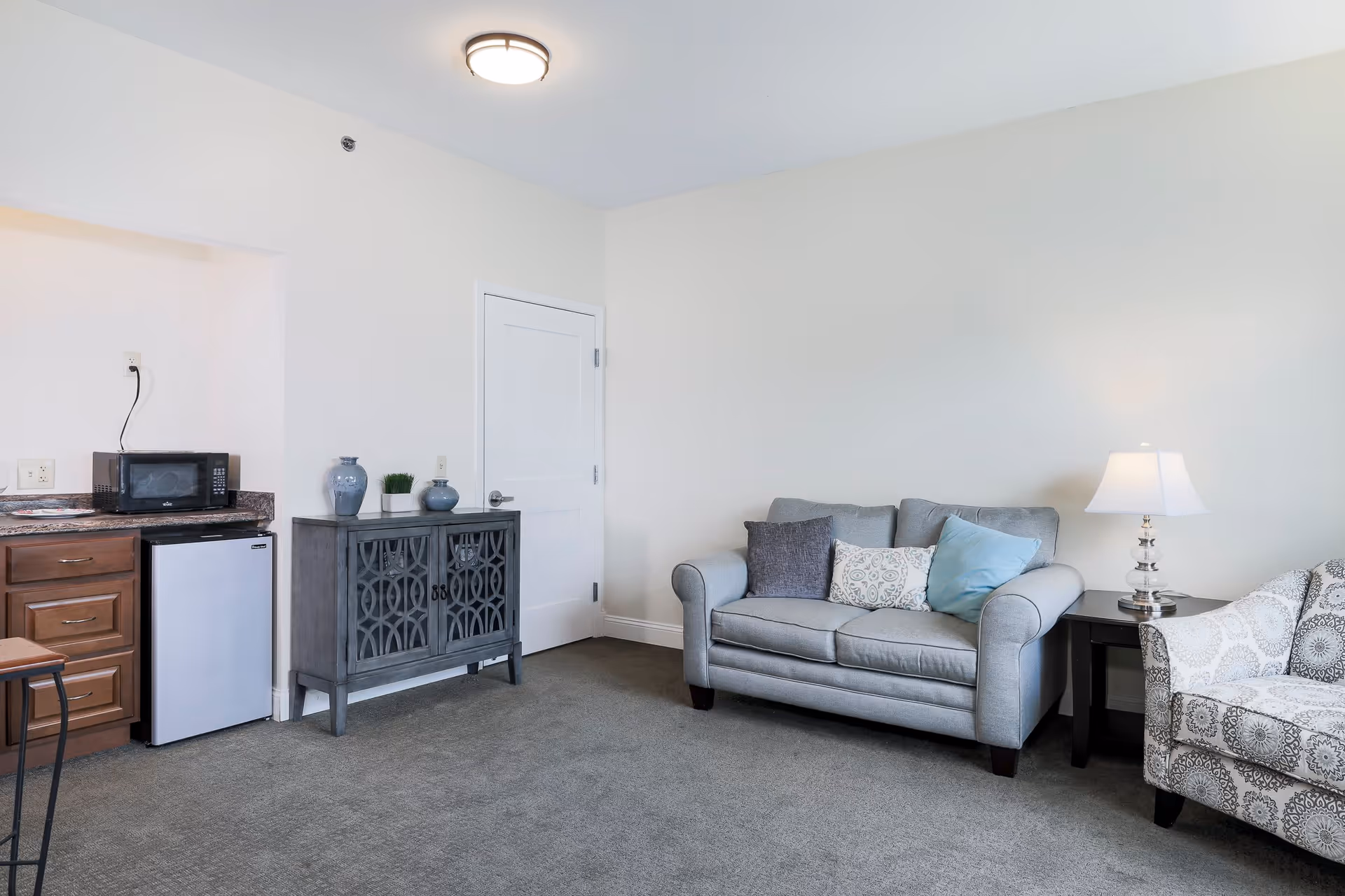 A cozy living room area with a gray loveseat adorned with three decorative pillows, a patterned armchair, and a side table with a lamp. To the left, there is a small kitchenette area with a microwave on the counter, a mini fridge below, and a gray cabinet with decorative vases and a small plant on top. The walls are painted white and the floor is carpeted in gray.