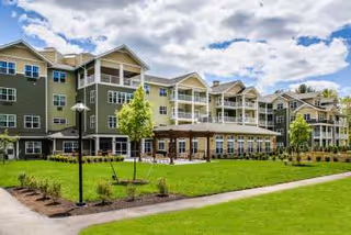 Exterior view of a multi-story senior living facility with green siding and white trim, featuring balconies and large windows. In front of the building is a well-maintained lawn with young trees, a paved walkway, and a wooden pergola structure. The sky is partly cloudy.