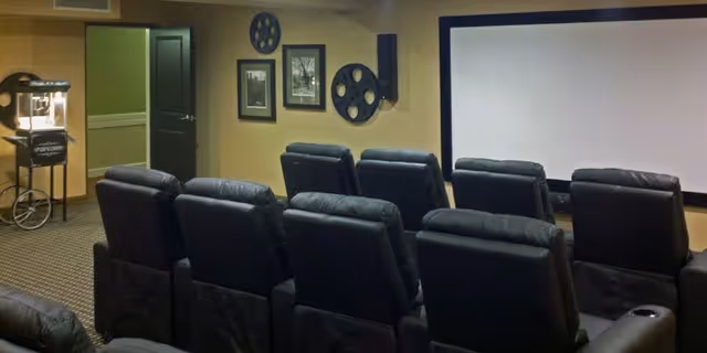 A small theater room with two rows of black leather recliner chairs facing a large white projection screen. The walls are decorated with framed pictures and large film reel decorations. A popcorn machine is visible in the corner near an open door.