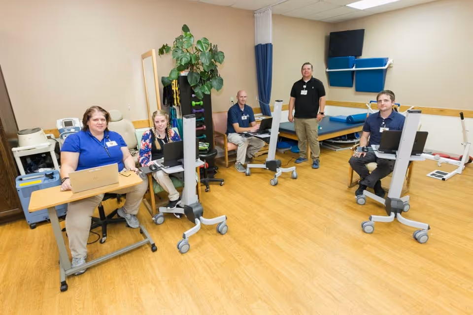 A group of five healthcare workers in a rehabilitation room with medical equipment, laptops, and exercise mats. The room has wooden flooring, beige walls, and a potted plant. Some workers are seated while others stand, all wearing ID badges.