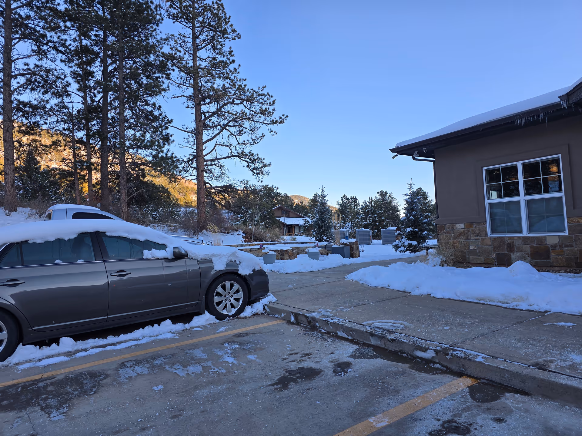 Snow-covered parking lot with a gray car partially covered in snow parked next to a building with stone and stucco exterior. Snow blankets the ground and trees in the background under a clear blue sky.