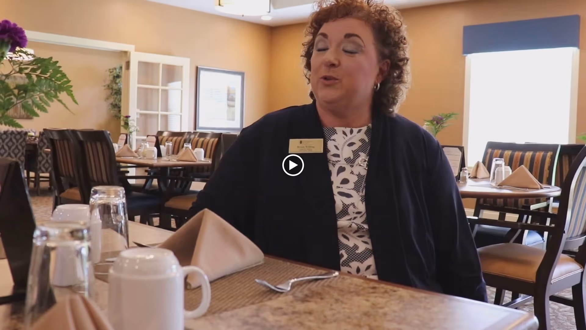 A woman seated at a set dining table in a dining room with neatly folded napkins and place settings.