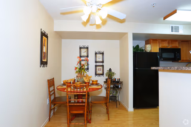 Dining area with a round wooden table set for four with plates, napkins, and a floral centerpiece. Four wooden chairs surround the table. The walls are decorated with framed artwork. To the right is a kitchen area with wooden cabinets and a black refrigerator. A ceiling fan with lights is above the dining table.