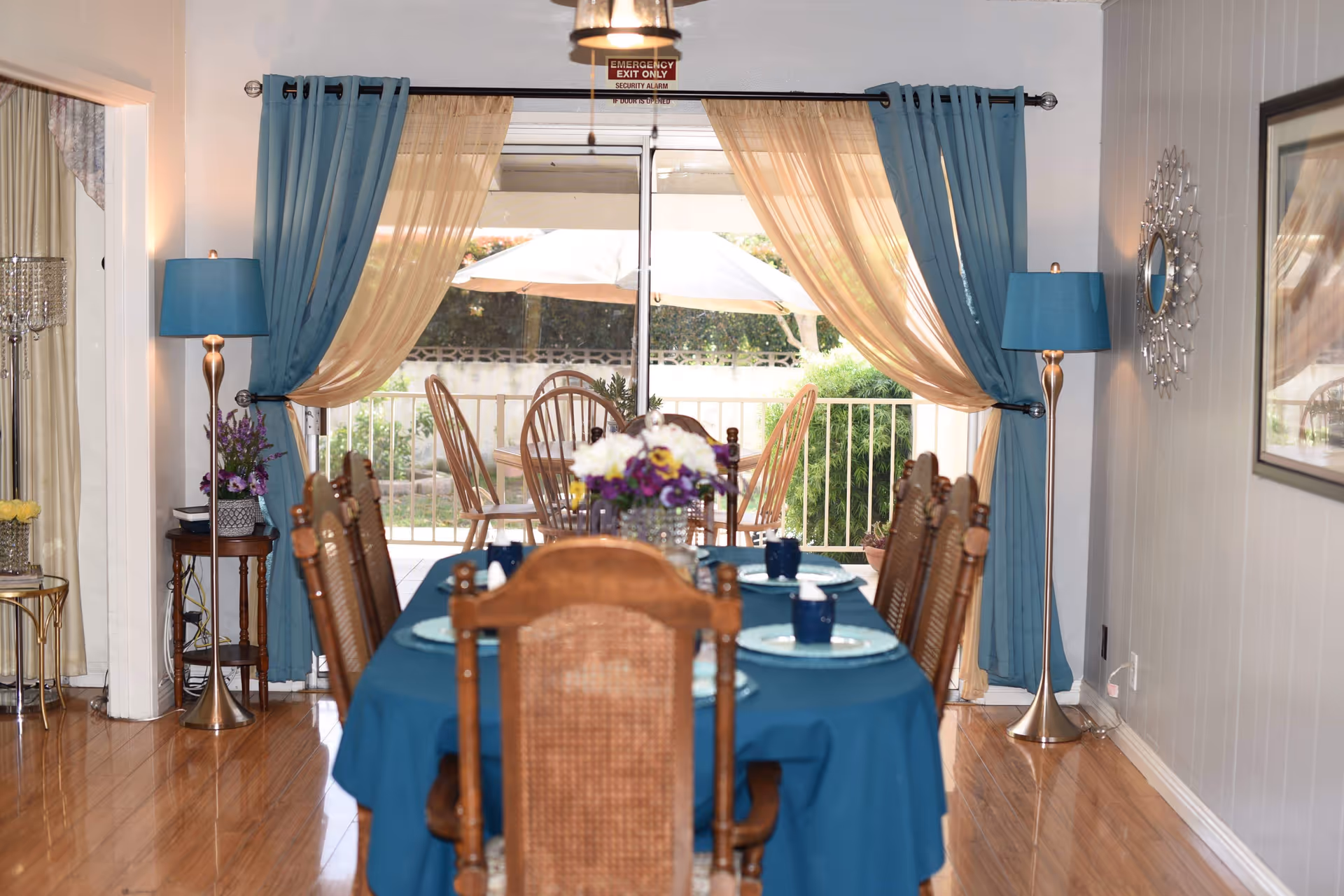 A dining room with a long table covered in a blue tablecloth and place settings, wooden chairs, floor lamps, and blue-and-gold curtains framing a sliding glass door to an outdoor patio.