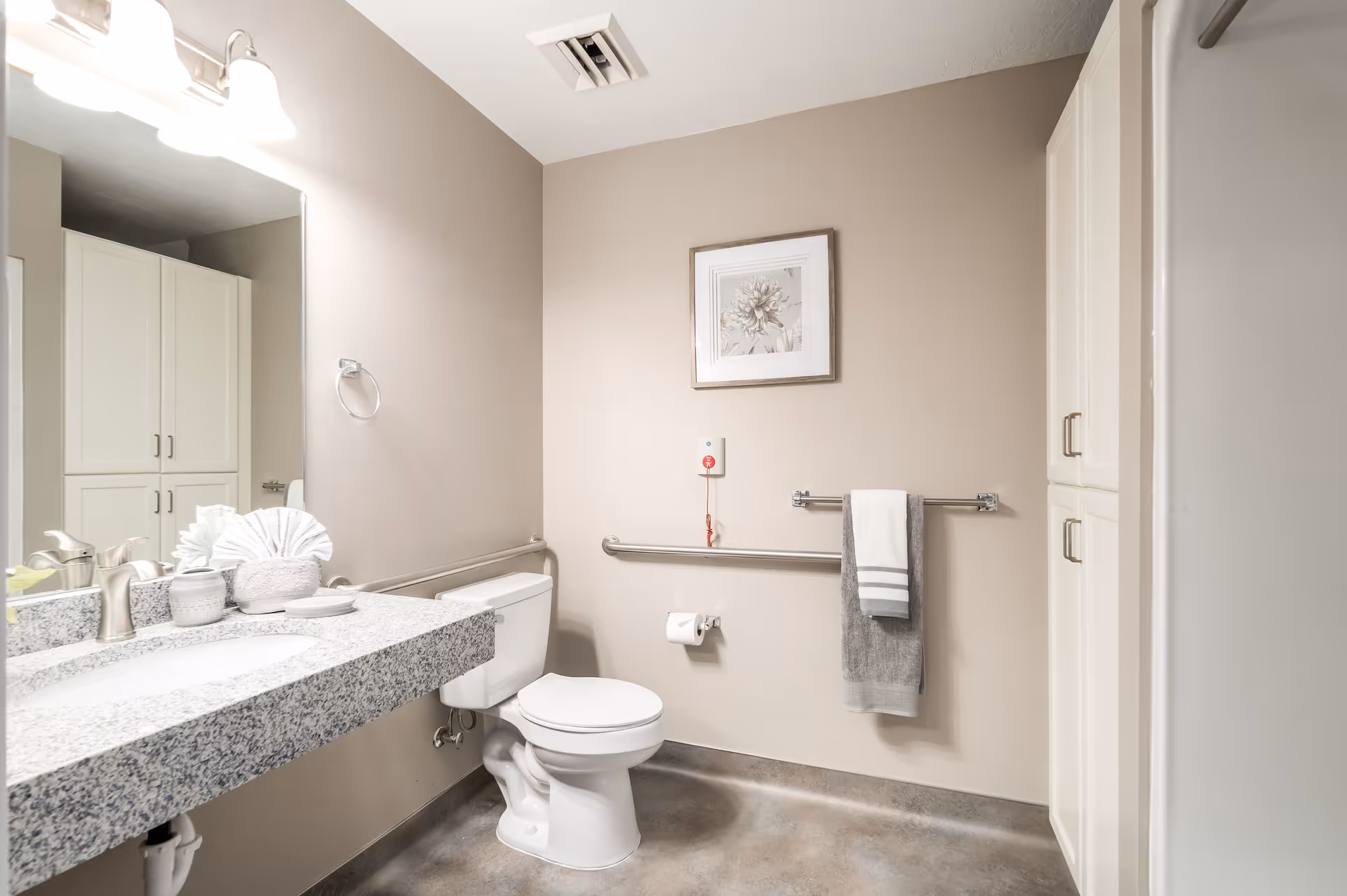 A clean and accessible bathroom with a white toilet, a granite countertop with a sink, a large mirror, and a cabinet. The walls are beige, and there is a framed flower picture above the toilet. A towel rack with two towels and a call button with a red cord are mounted on the wall.