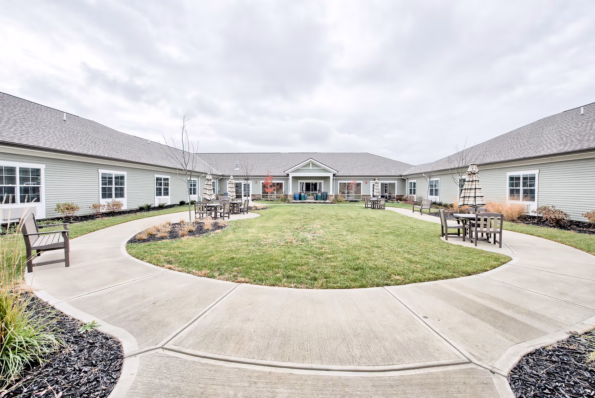 Outdoor courtyard area of a senior living facility with a circular concrete walkway surrounding a grassy lawn. There are several wooden benches and tables with closed umbrellas placed along the walkway. The building surrounds the courtyard with multiple windows and a covered entrance in the center. The sky is overcast.
