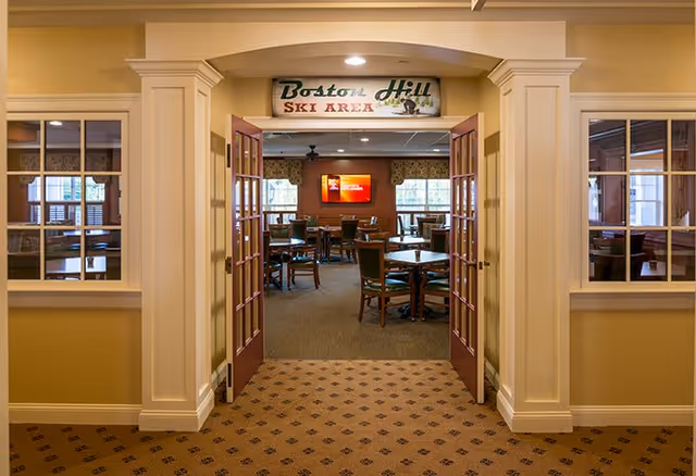 View through an open double door into a room with multiple tables and chairs arranged for dining or socializing. Above the door is a sign that reads 'Boston Hill Ski Area'. The room has large windows with curtains and a television mounted on the far wall.