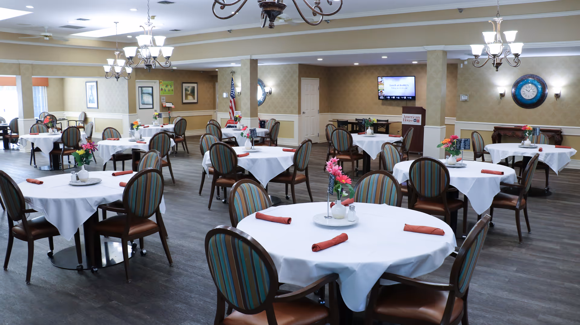 A spacious dining room with multiple round tables covered with white tablecloths, each set with rolled red napkins and small flower arrangements. The room has wooden flooring, beige patterned walls, chandeliers hanging from the ceiling, and a television mounted on the wall displaying a message about lunch. There is an American flag in the corner and a podium with the American House logo.