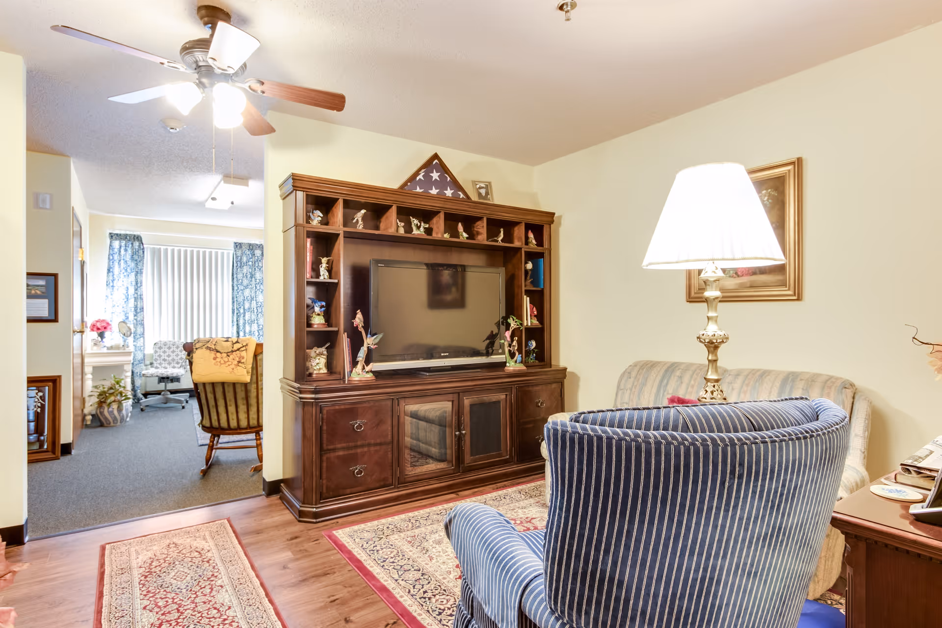 A cozy living room area with a wooden entertainment center holding a flat-screen TV and decorative figurines. In front of the entertainment center is a striped blue armchair and a beige sofa with a table lamp on a side table. The room has hardwood flooring with area rugs and a ceiling fan with lights. In the background, there is an adjoining room with a window covered by vertical blinds and blue patterned curtains, a desk, and a rocking chair.