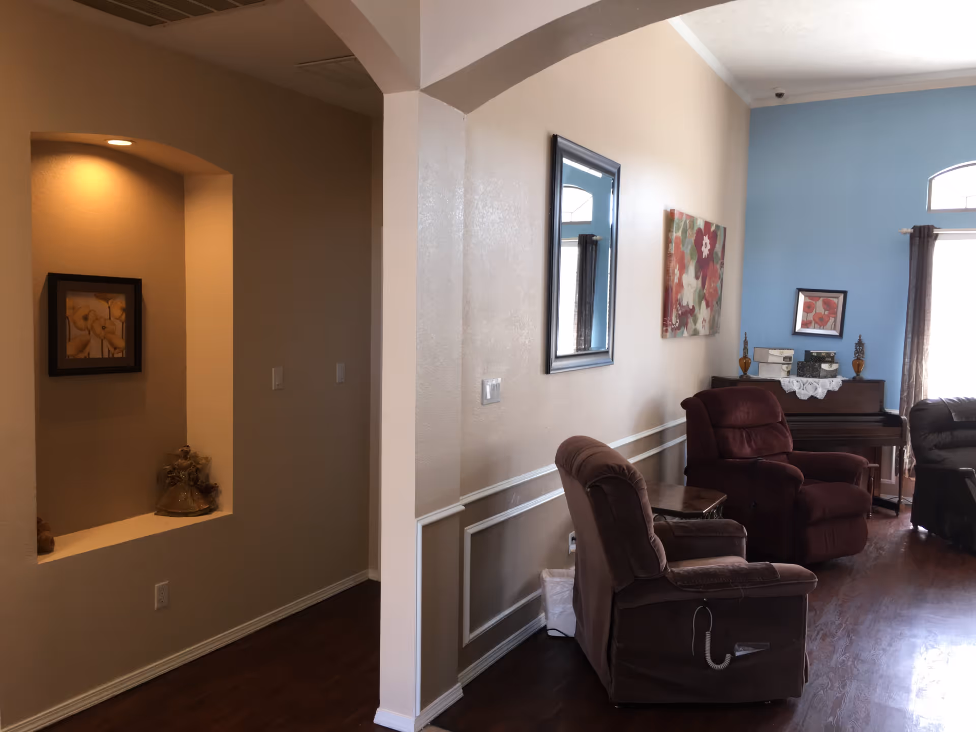 Interior view of a living room area with two recliner chairs, a wooden side table, a piano against a blue accent wall, and framed artwork on the walls. There is a small alcove with a light and decorative items on the left side.