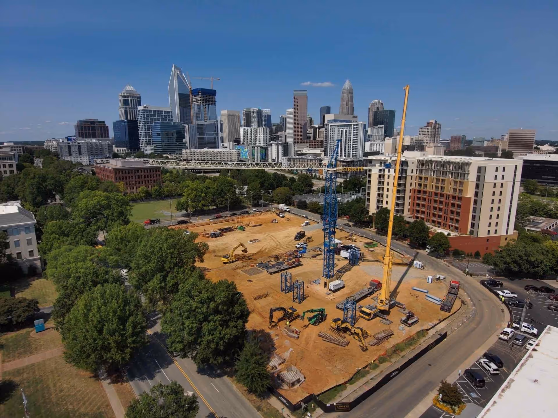 Aerial view of a construction site with cranes and construction equipment, surrounded by trees and roads, with a city skyline in the background under a clear blue sky.