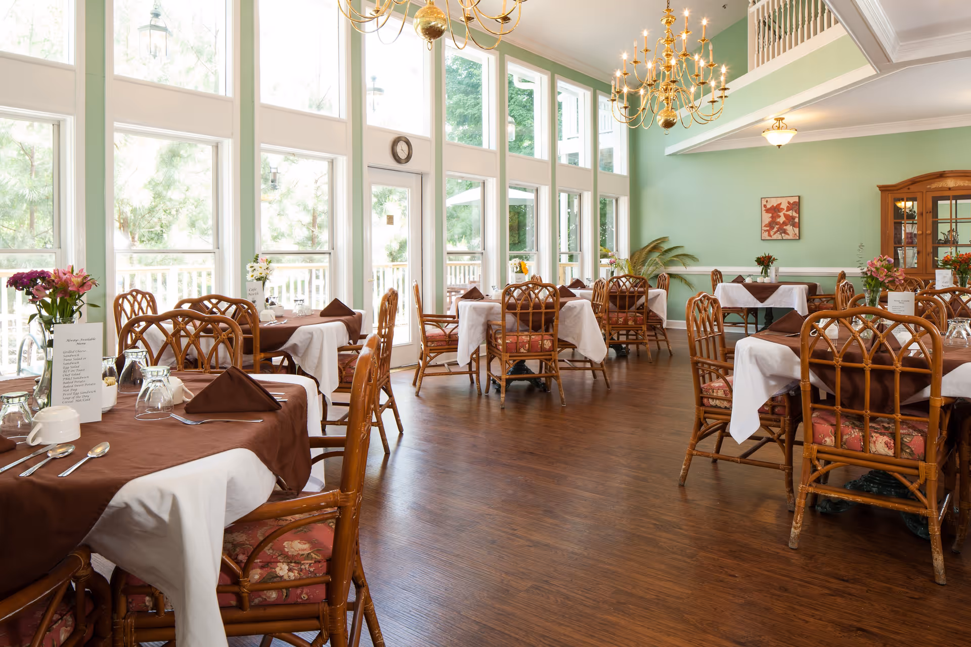 Sunlit dining room with multiple set tables, wicker chairs, large windows and chandeliers.