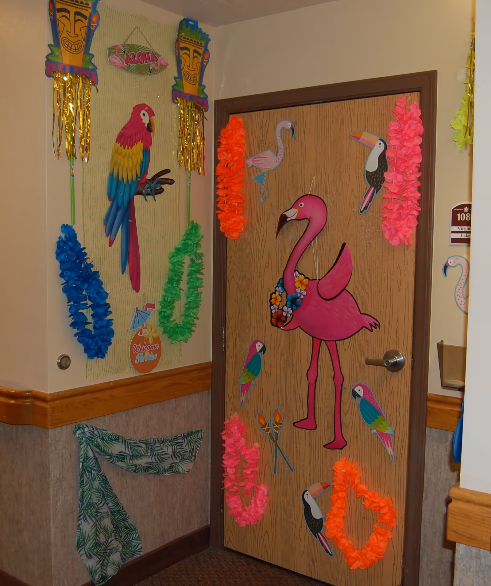 A decorated door and adjacent wall in a hallway with a tropical theme. The door features a large pink flamingo wearing a flower lei, surrounded by smaller images of flamingos, toucans, and parrots. The wall has colorful leis hanging and tiki-themed decorations, including a parrot and a sign that says 'Welcome Aloha'.