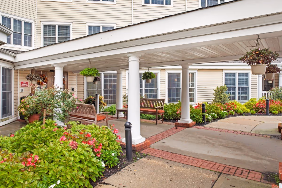 Covered entrance patio with white columns, benches, hanging plants and landscaped flower beds in front of a multi‑window building.