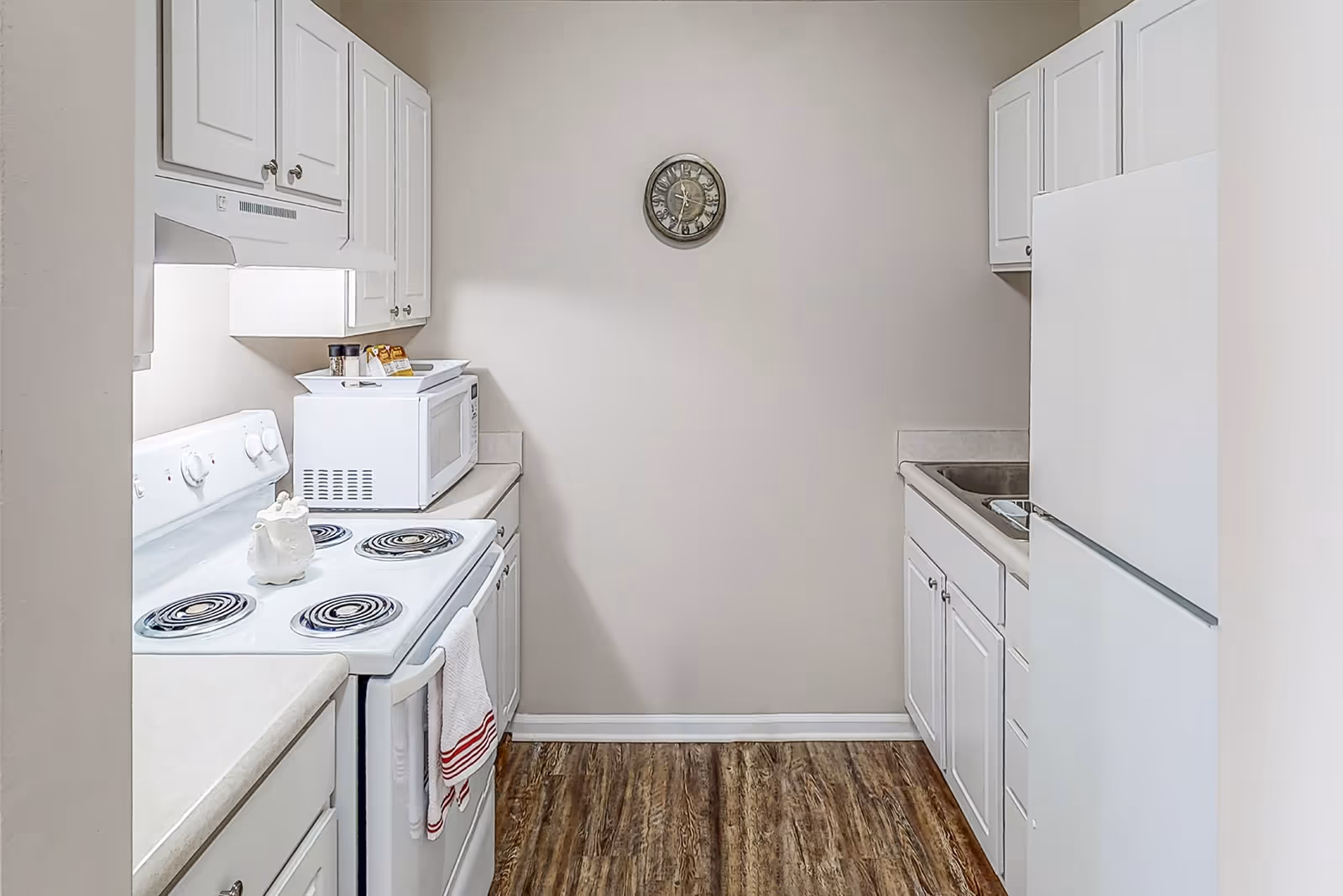A small kitchen with white cabinets, a white electric stove with coil burners, a white microwave on the counter, a white refrigerator, and a stainless steel sink. There is a clock on the beige wall and wood-patterned flooring.