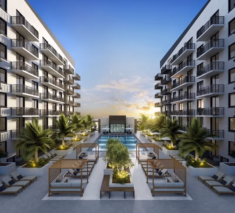 Outdoor courtyard area of a modern residential or senior living facility at sunset, featuring a central swimming pool surrounded by palm trees, lounge chairs, and cabanas with cushioned seating. Two multi-story buildings with balconies flank the pool area.