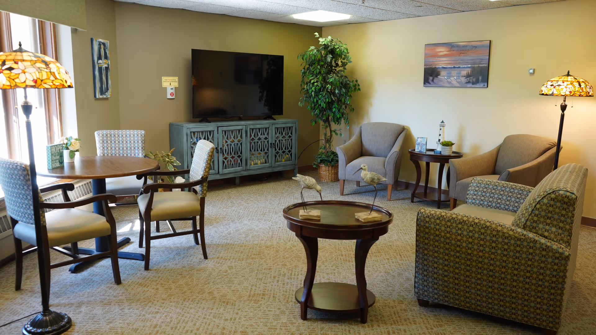 A cozy living room area with a round wooden table and four upholstered chairs near a window on the left. In the center, there is a round wooden coffee table with decorative bird figurines. The back wall features a flat-screen TV on a blue cabinet, a potted plant, and two armchairs with a small side table between them. The room is softly lit by two Tiffany-style floor lamps and has a carpeted floor and beige walls with framed artwork.