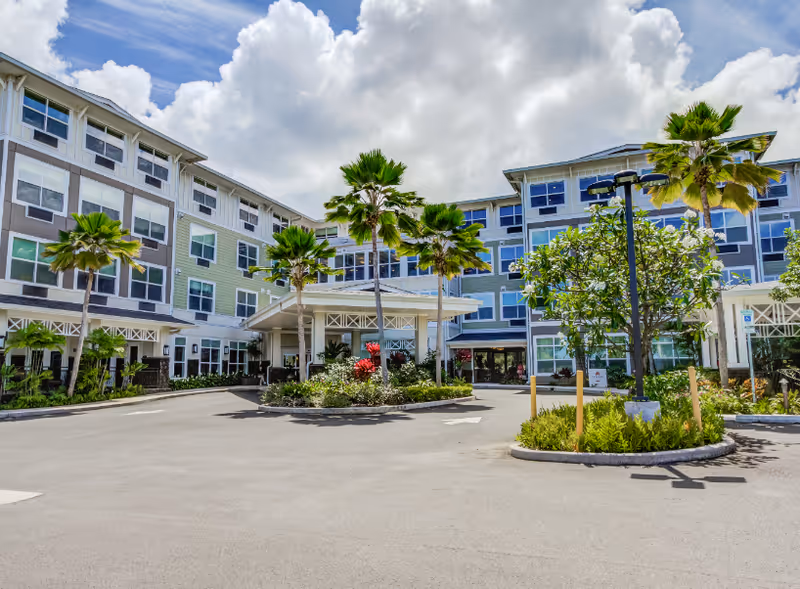 Exterior view of The Plaza at Kaneohe senior living facility showing a multi-story building with numerous windows, palm trees, landscaped greenery, and a covered entrance under a partly cloudy sky.