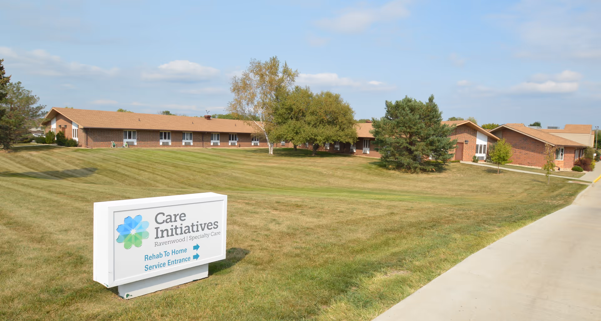 Exterior view of a single-story brick building with a brown roof, surrounded by a well-maintained lawn and trees under a partly cloudy sky. A white sign in the foreground reads 'Care Initiatives Ravenwood Specialty Care Rehab To Home Service Entrance' with arrows pointing to the right.