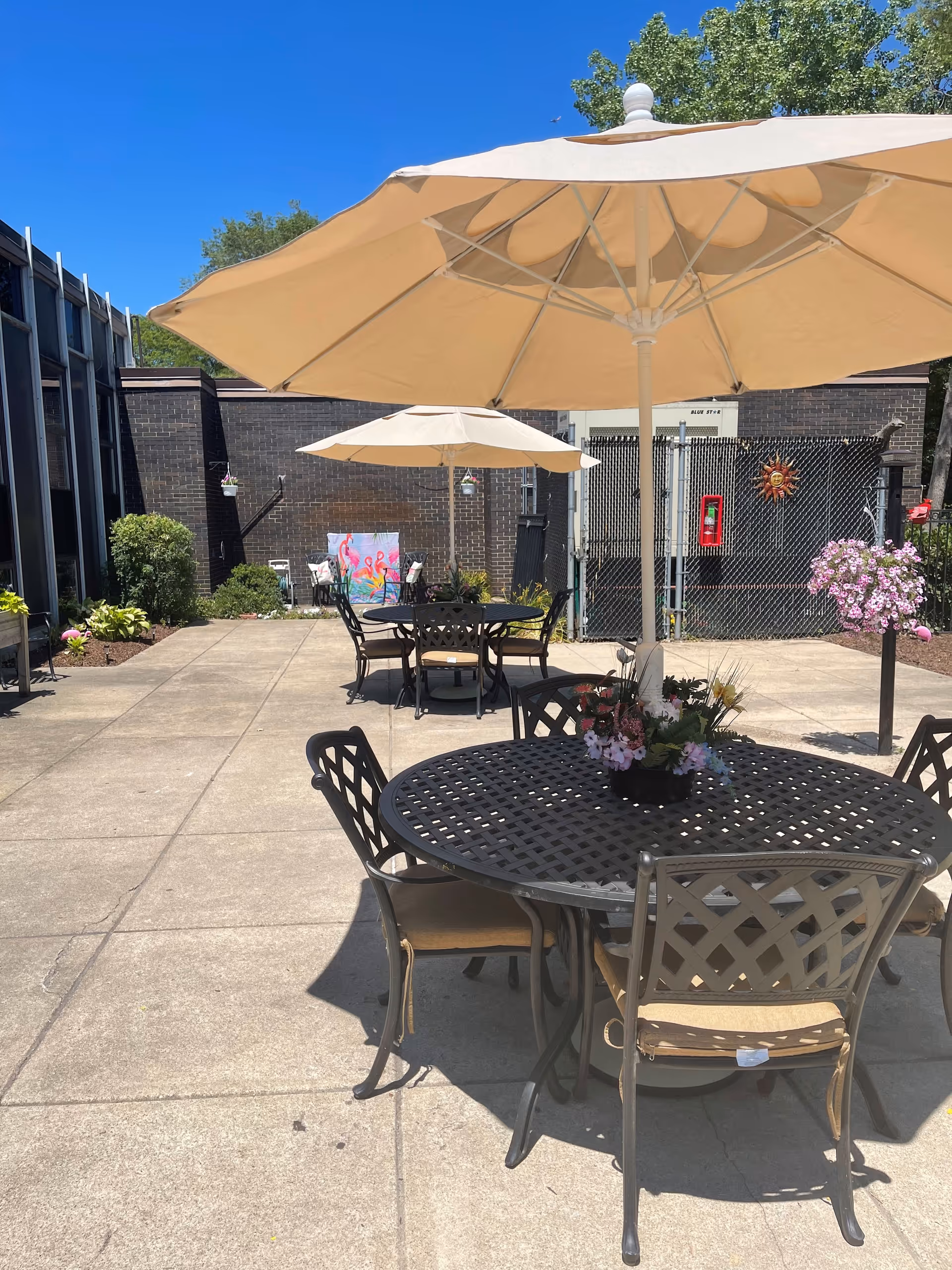 Outdoor patio area with round metal tables and chairs, each table shaded by large beige umbrellas. There are flower arrangements on the tables and hanging flower baskets nearby. The patio is surrounded by a brick building and a chain-link fence, with some greenery and a bright blue sky overhead.