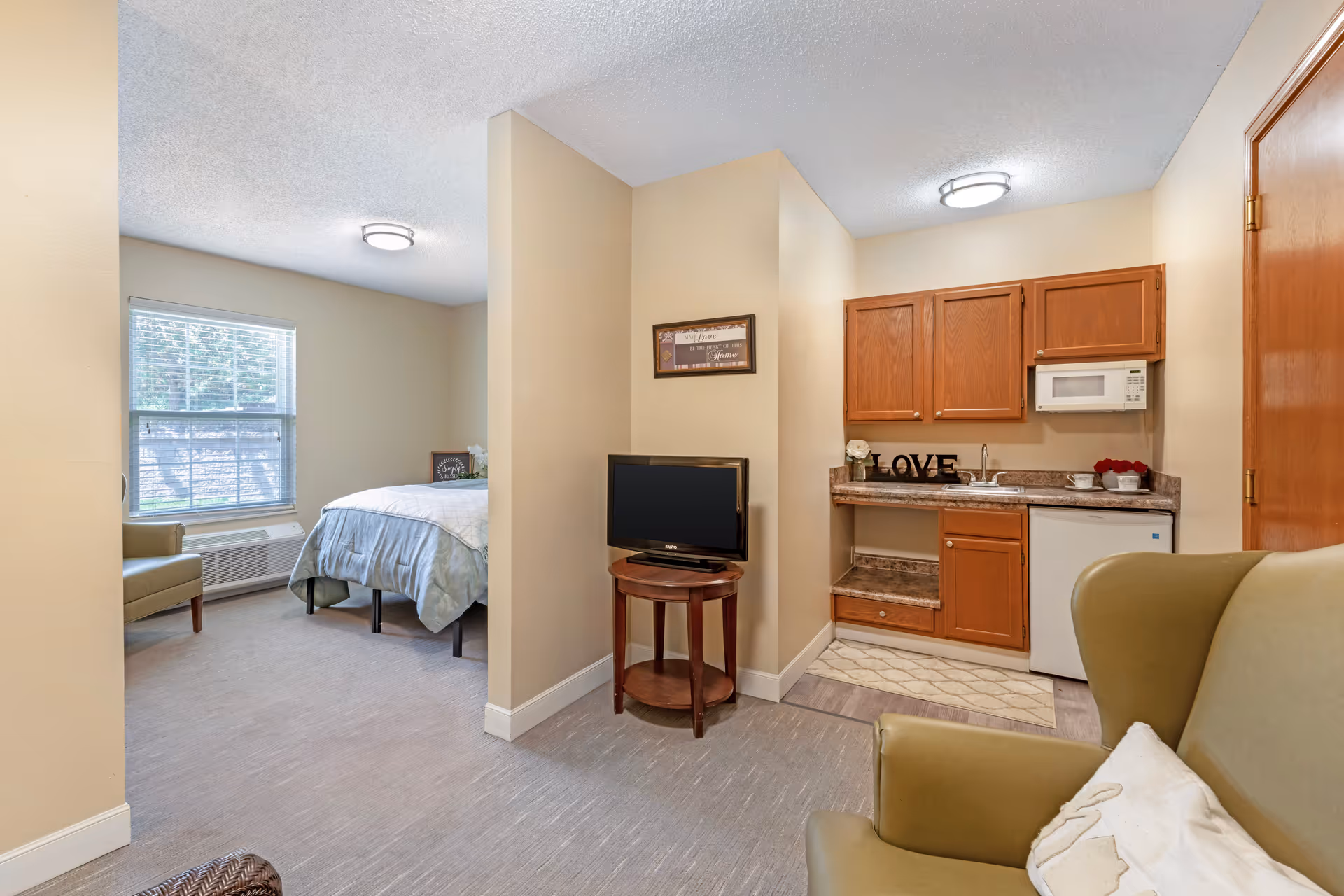 Studio-style senior living interior showing a bed area, small kitchenette with cabinets and microwave, a TV on a stand, and seating.