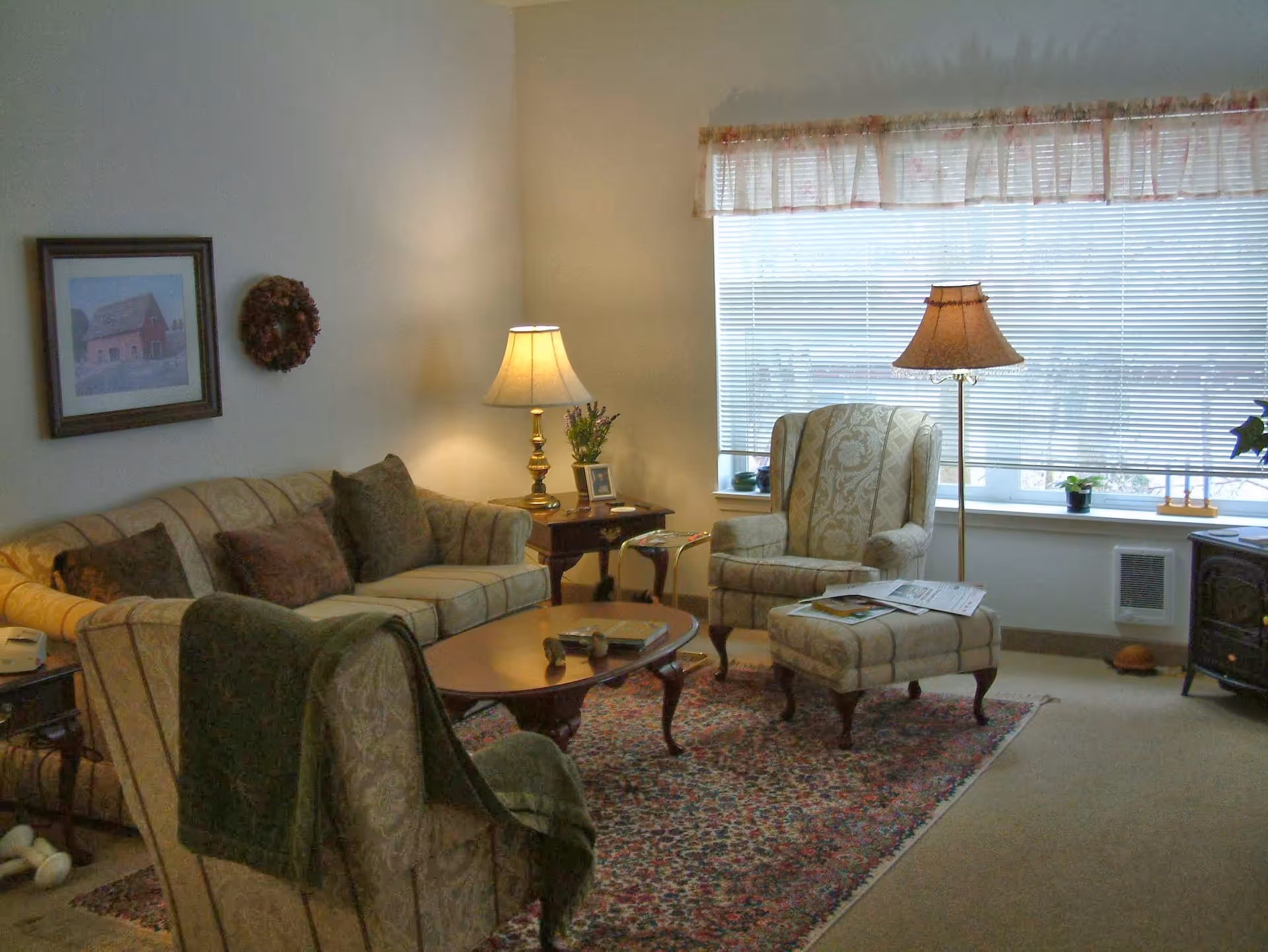 Cozy living room with a sofa, armchairs, ottoman, coffee table, lamps and a large window with blinds.