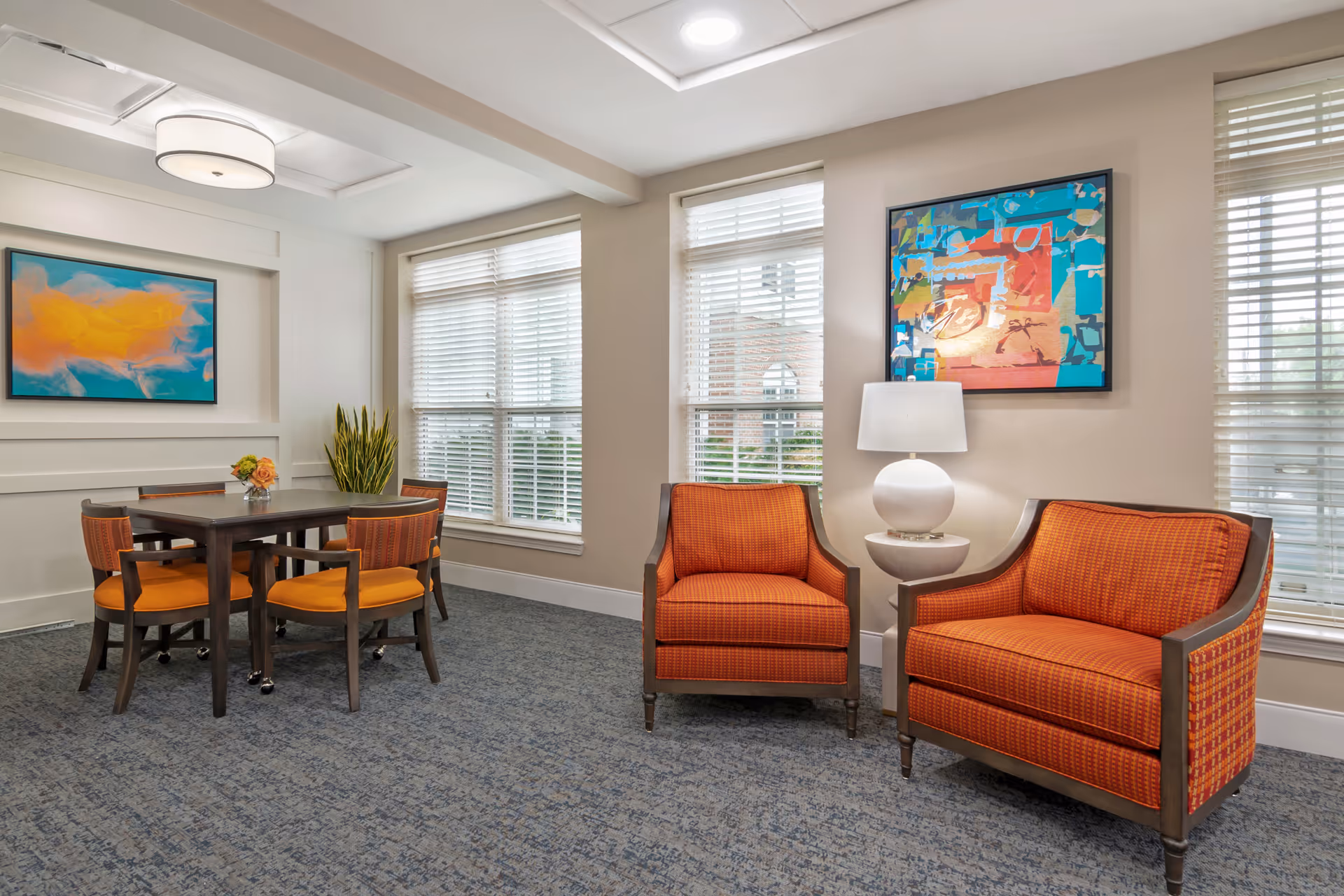 Bright common room with two orange upholstered armchairs, a round side table lamp, and a dining table with matching chairs by large windows.