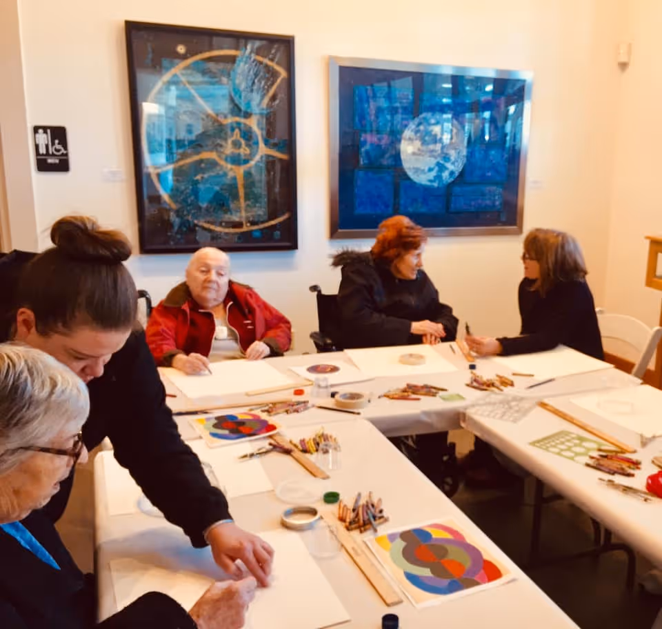 A group of elderly people and a caregiver engaged in an art activity around tables covered with white paper, colored pencils, and art supplies in a well-lit room with abstract paintings on the walls.