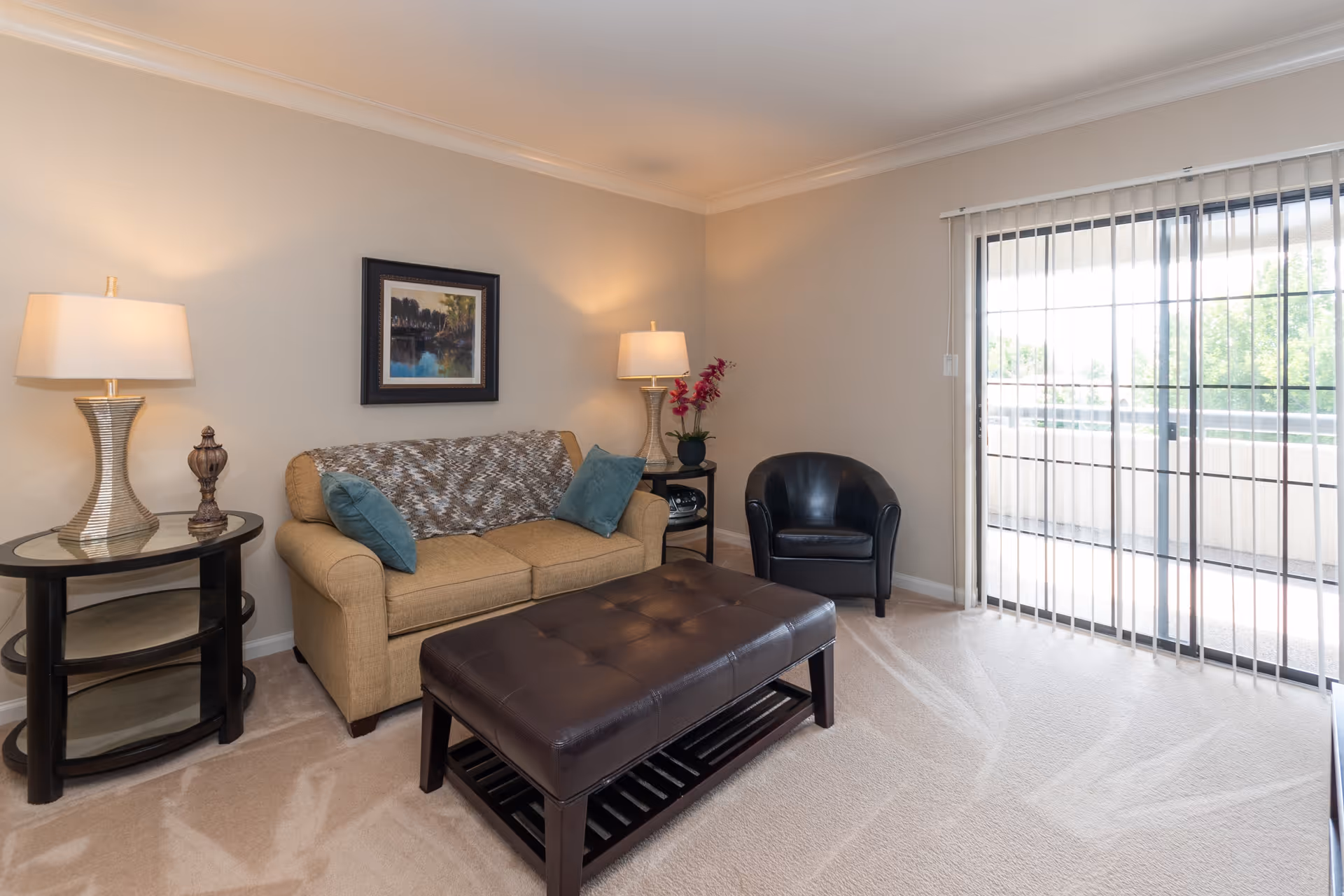 Bright living room with a sofa, armchair, leather ottoman coffee table, side tables with lamps, wall art, and sliding glass doors with vertical blinds.