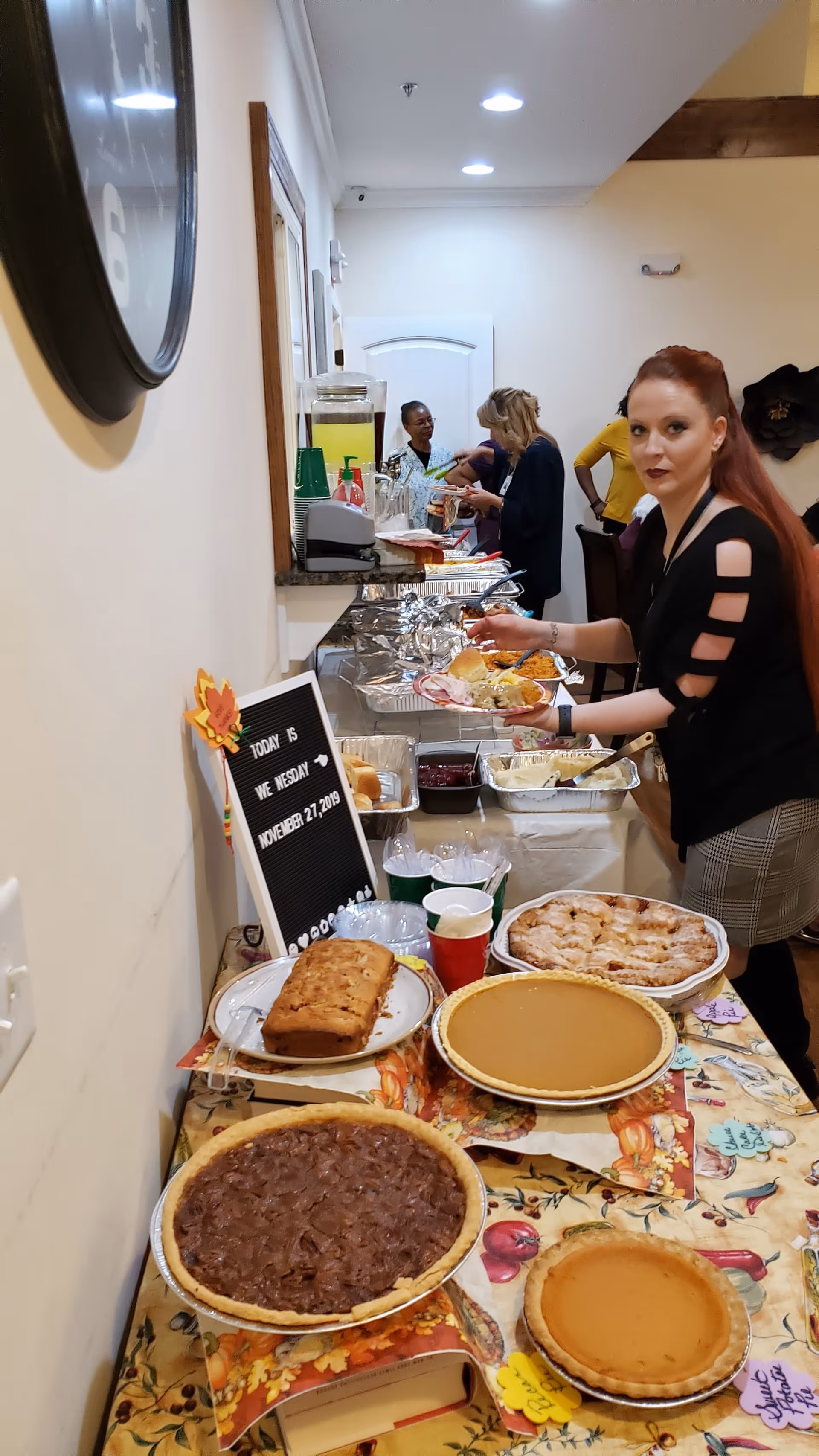 A buffet table set up with various desserts including pecan pie, pumpkin pies, and a loaf cake, along with a sign displaying the date Wednesday, November 27, 2019. A woman with long red hair is serving herself food from trays of hot dishes further down the table. Several other people are visible in the background near the serving area in a well-lit indoor space.