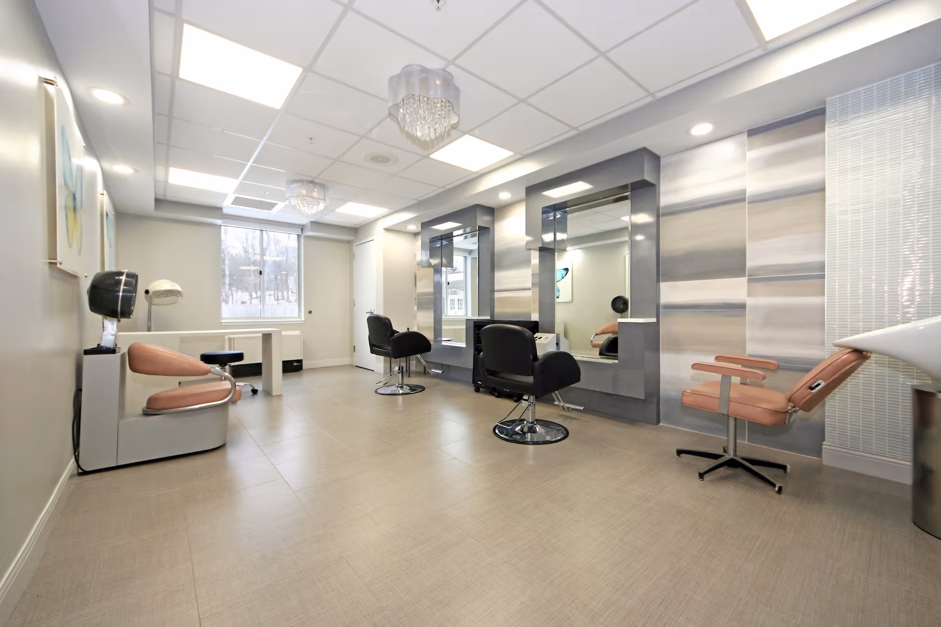 A bright and modern hair salon area inside a nursing and rehabilitation center, featuring salon chairs in front of large mirrors, a hair dryer chair, and a wash basin. The room has tiled flooring, recessed lighting, and a window letting in natural light.