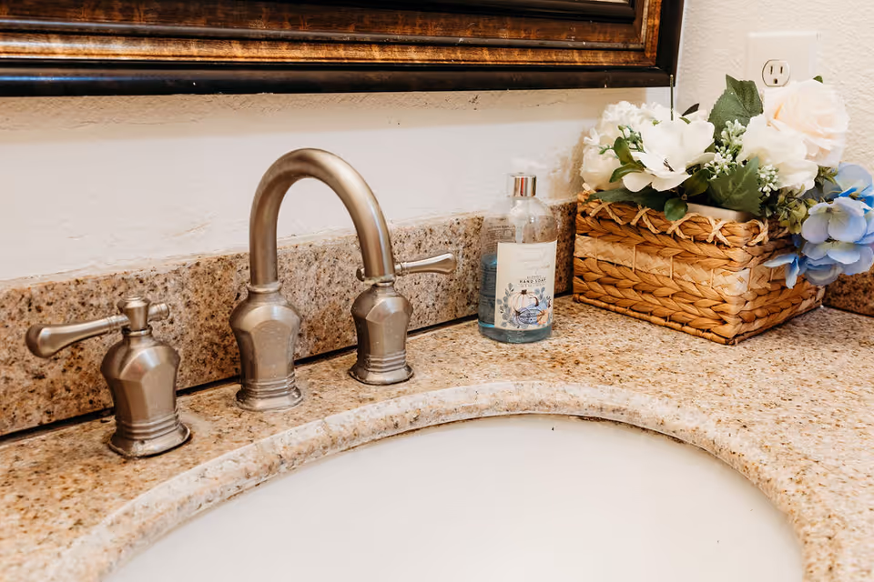 Close-up view of a bathroom sink with a beige granite countertop, a silver faucet with two handles, a bottle of hand soap, and a woven basket containing white and blue artificial flowers. A large mirror with a dark wooden frame is partially visible above the sink.