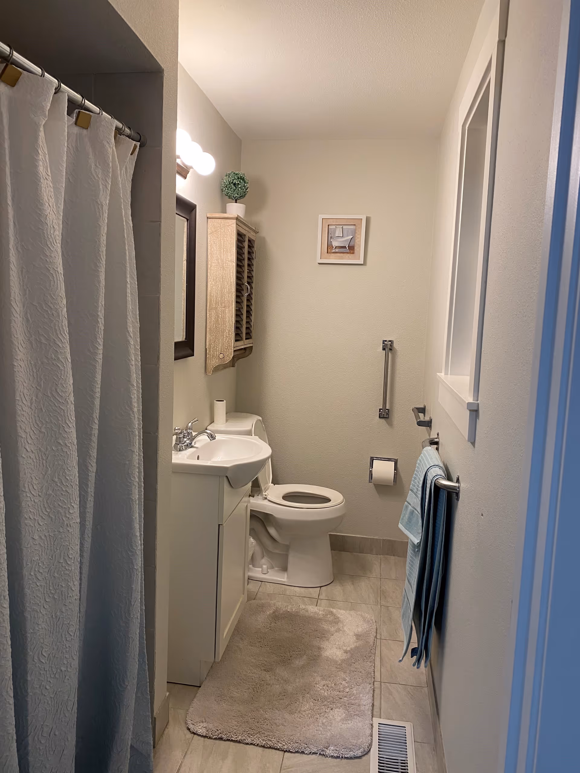 A small bathroom with a white toilet, a white sink with a cabinet, a wall-mounted cabinet with a small potted plant on top, a framed picture on the wall, a towel rack with blue towels, a white shower curtain, and a beige bath mat on the tiled floor.
