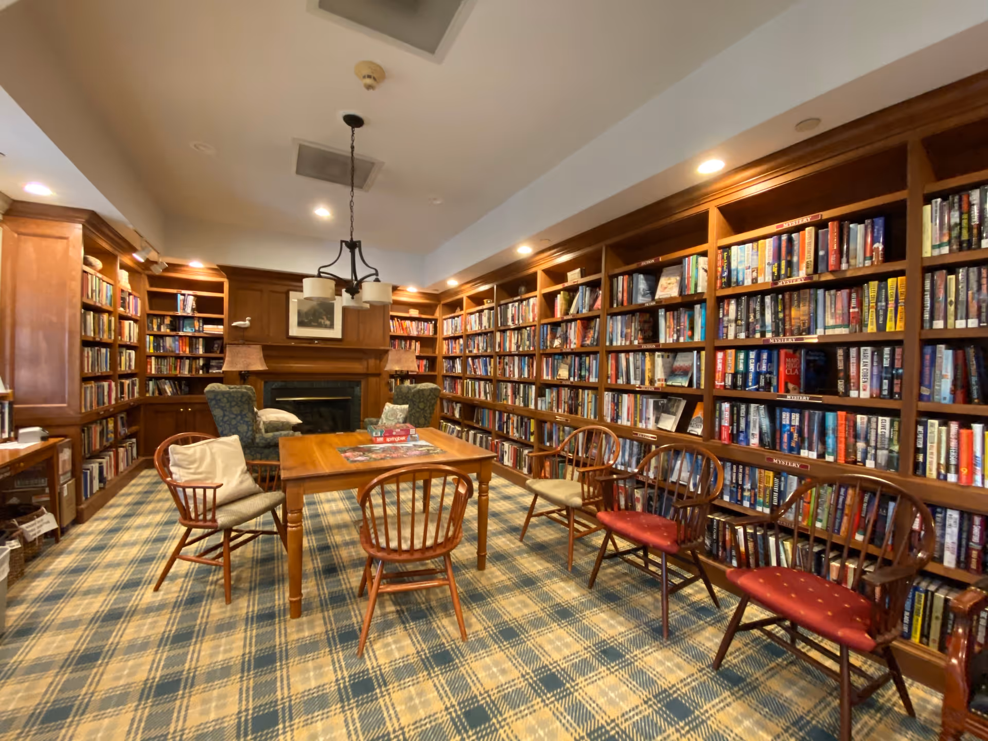 Cozy library room with wooden bookshelves lining the walls, a central table surrounded by chairs, patterned carpet, and a fireplace.