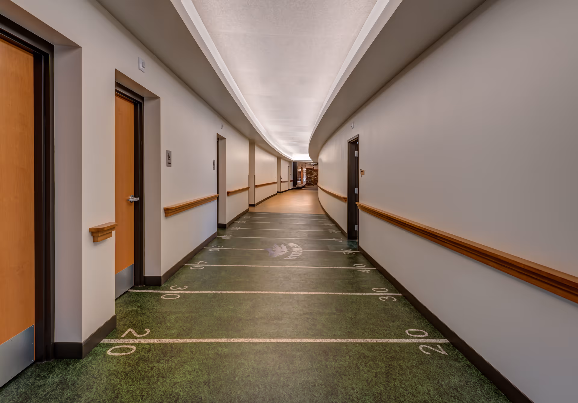 Long curved interior hallway with doors on each side, wooden handrails, and a green floor marked like a football field.