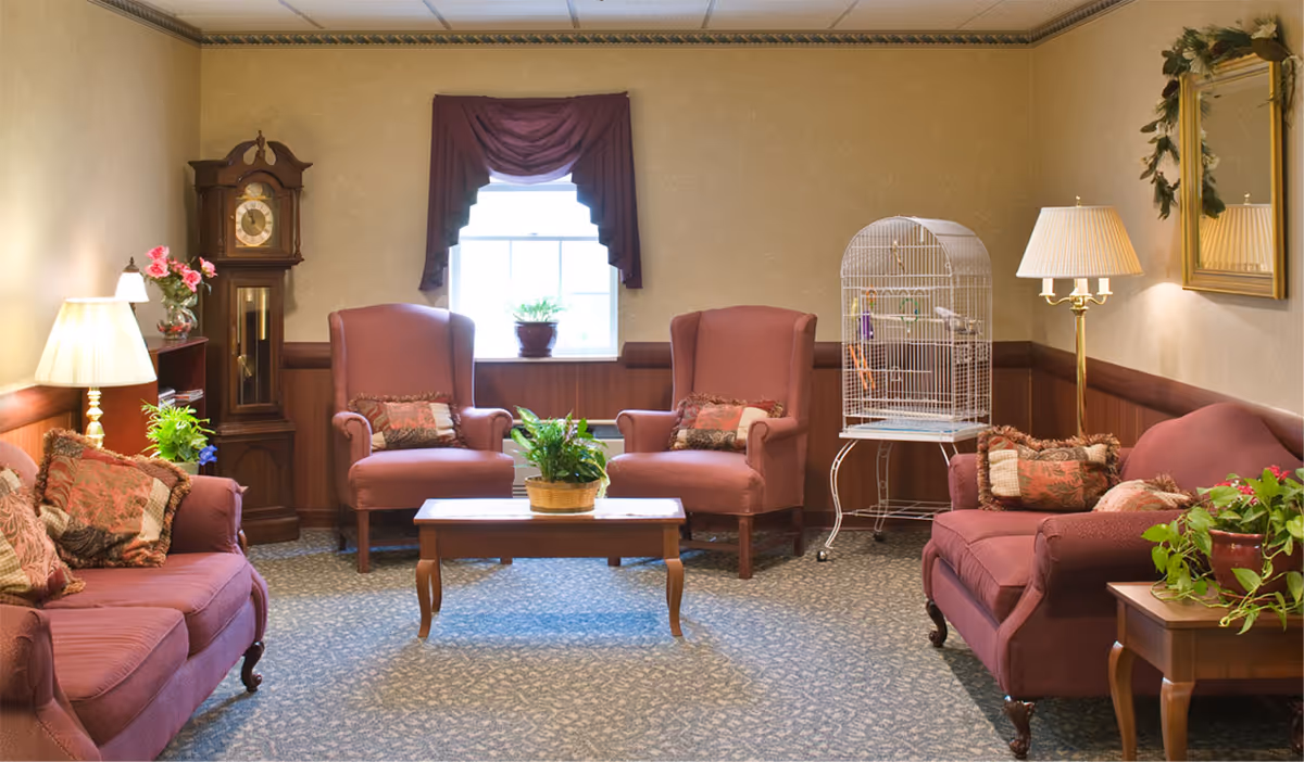 A cozy living room with two pink upholstered armchairs and two matching sofas arranged around a wooden coffee table with a potted plant. There is a tall grandfather clock in the corner, a birdcage on a stand, a floor lamp, and a side table with another potted plant. A window with a dark valance lets in natural light, and a mirror with a decorative wreath hangs on the wall.