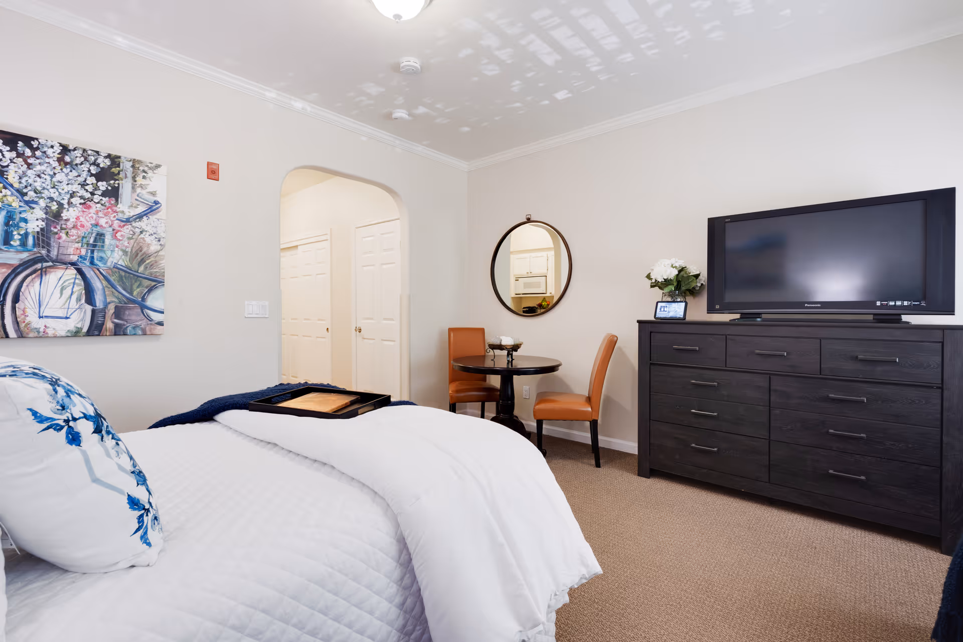 A cozy bedroom with a white quilted bedspread and blue floral pillows. There is a dark wooden dresser with a flat-screen TV on top, a small round table with two brown chairs, and a round mirror on the wall reflecting a kitchen area. A colorful painting of a bicycle with flowers hangs on the wall.