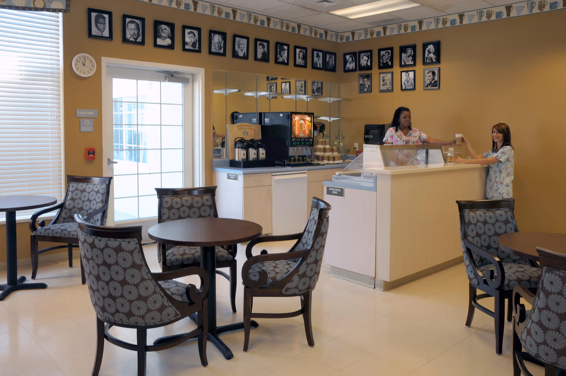 A small dining area with round tables and patterned chairs. Two women are interacting at a counter with a coffee and beverage station behind it. The walls are decorated with framed black and white portraits and a clock is mounted near a glass door with blinds.